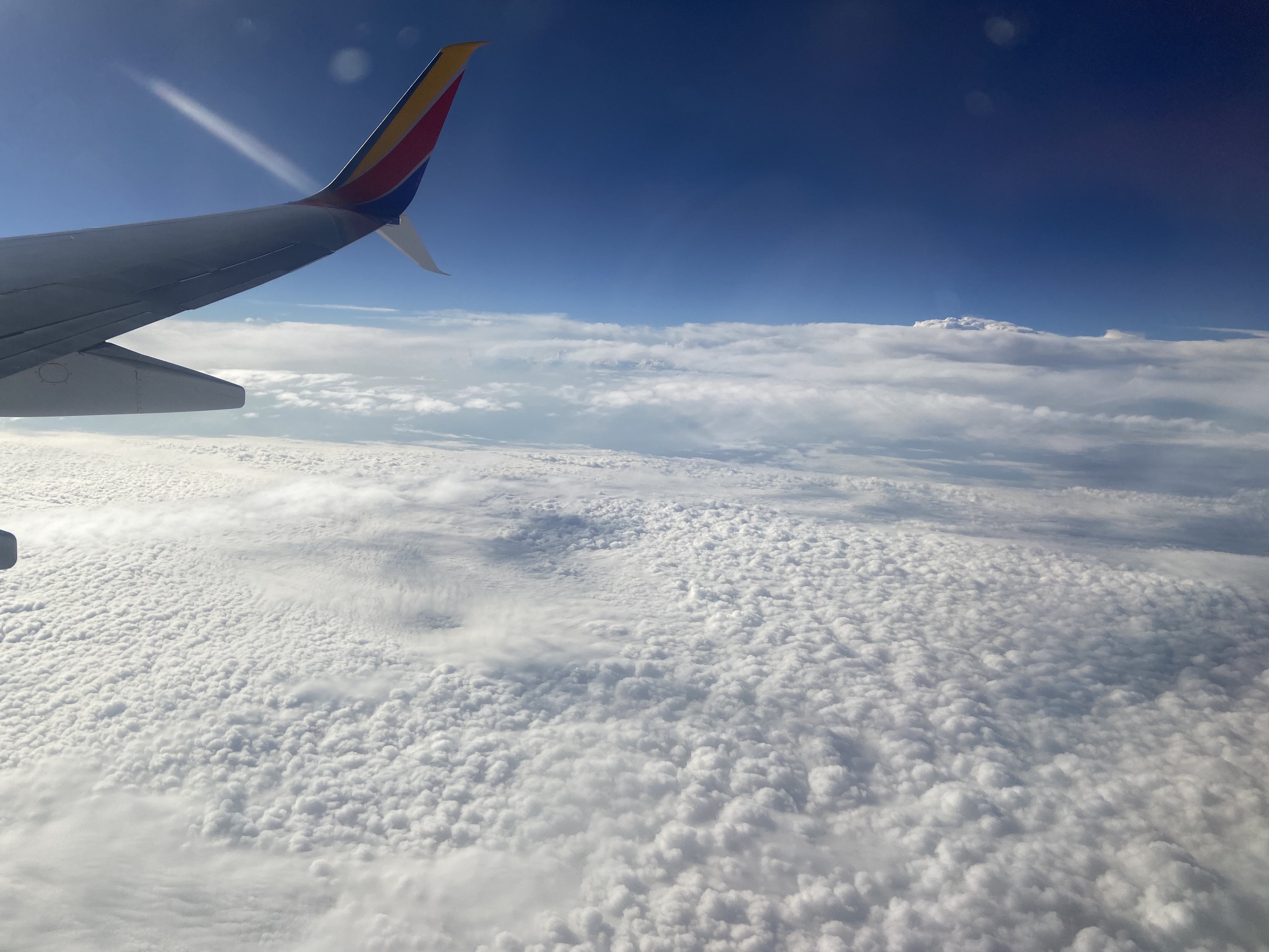 View from an airplane window showing the airplane wing and a vast landscape of clouds beneath a clear sky