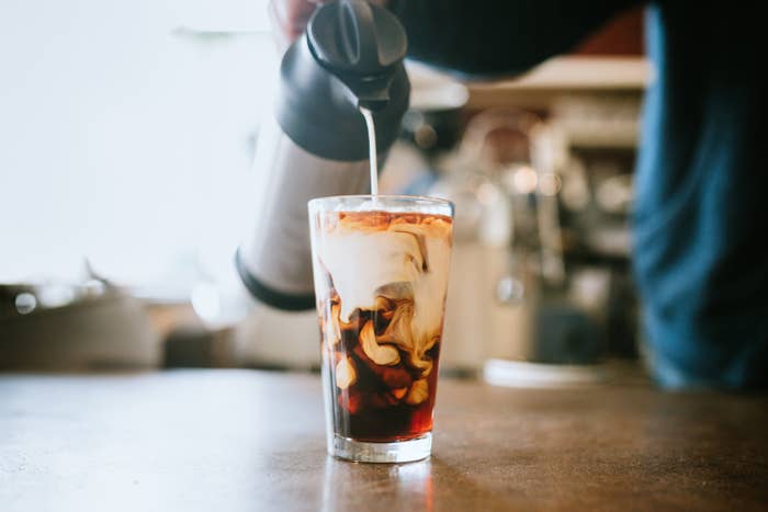 A close-up of someone pouring milk into a glass of iced coffee on a wooden table, creating a swirling effect