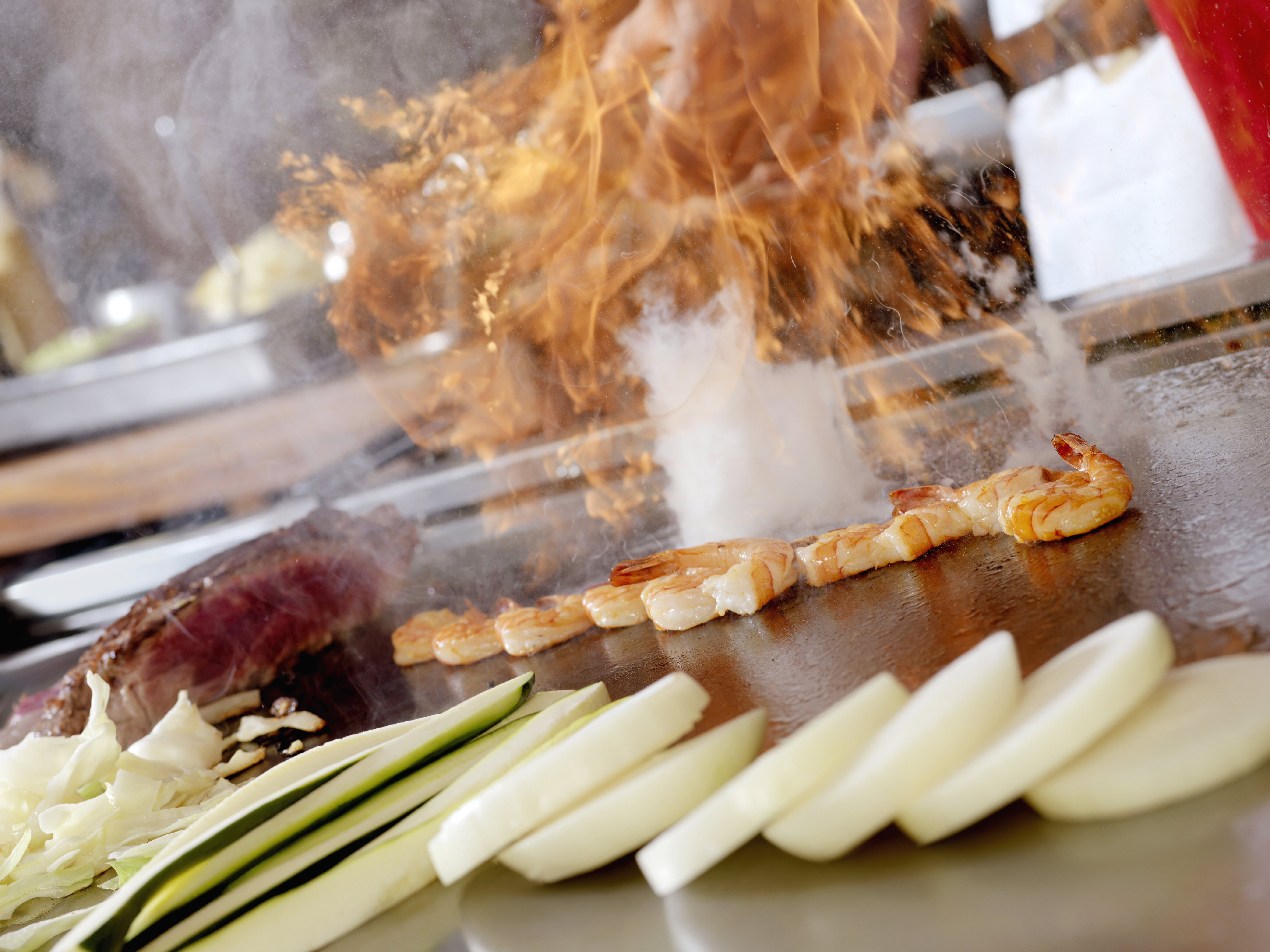 Shrimp and vegetables being cooked on a teppanyaki grill with flames and steam rising in the background