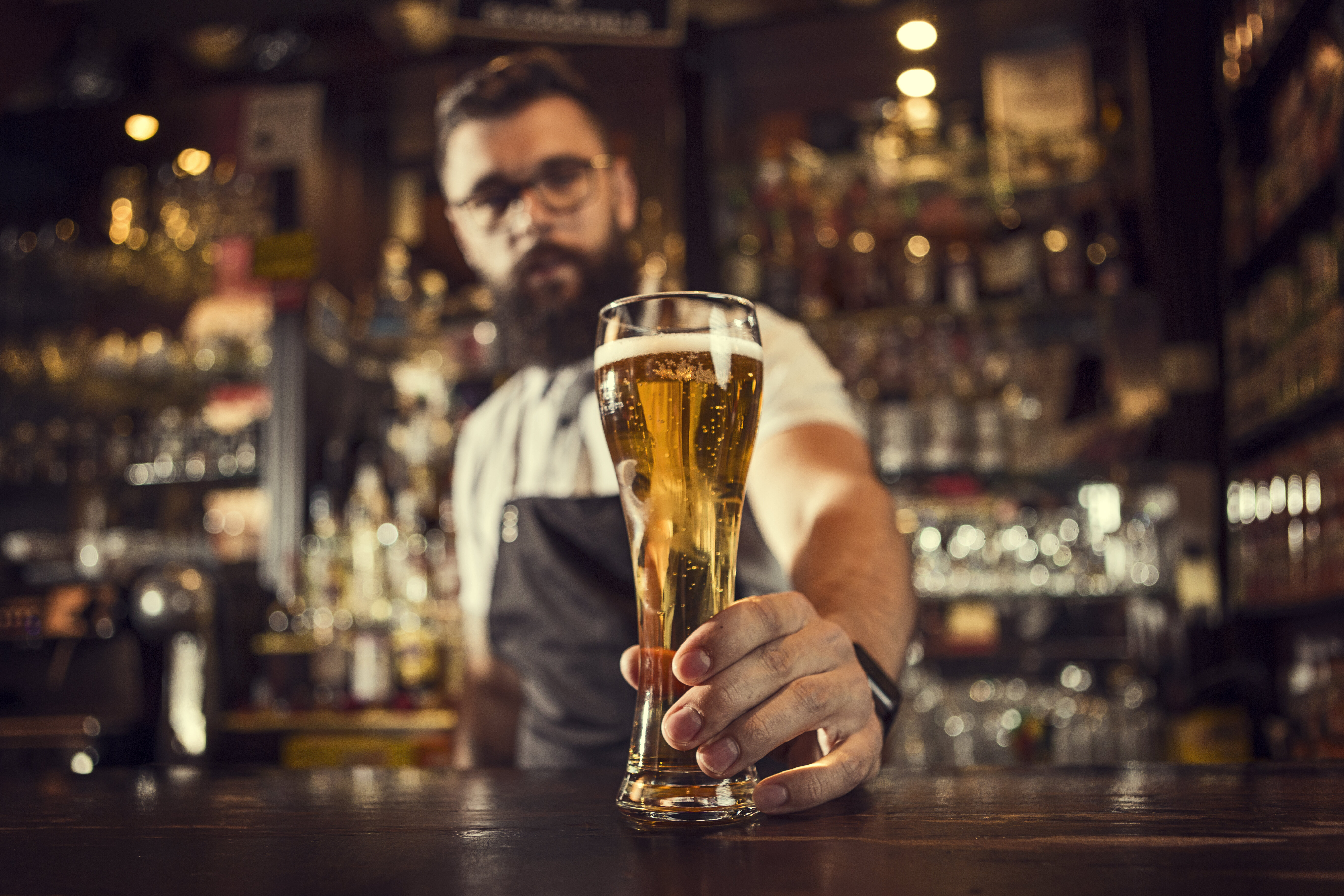 Bartender with a beard and glasses serves a tall glass of beer in a busy pub. Glasses and bottles line the shelves behind him