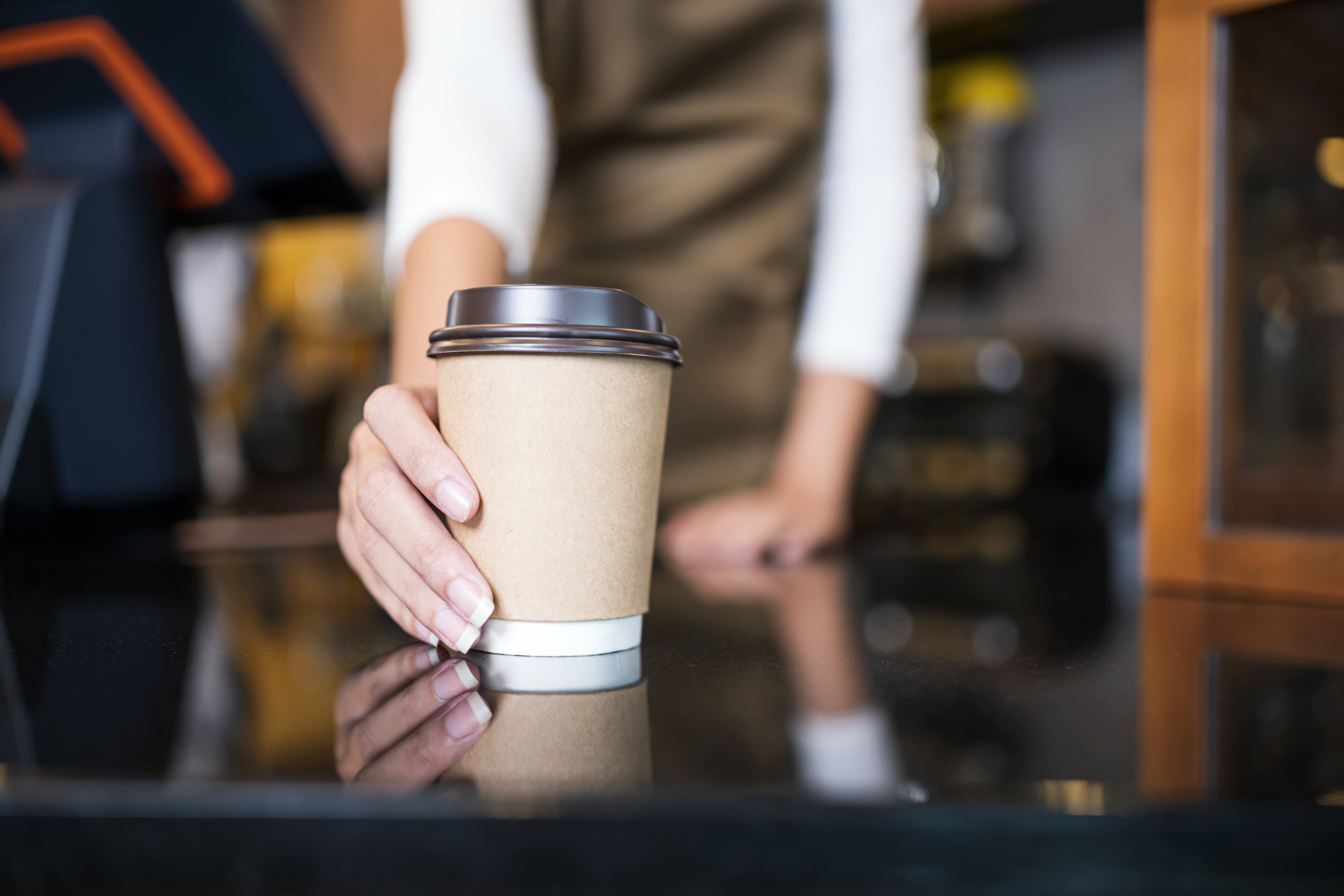 A person in a coffee shop extending a paper coffee cup with a lid over the counter