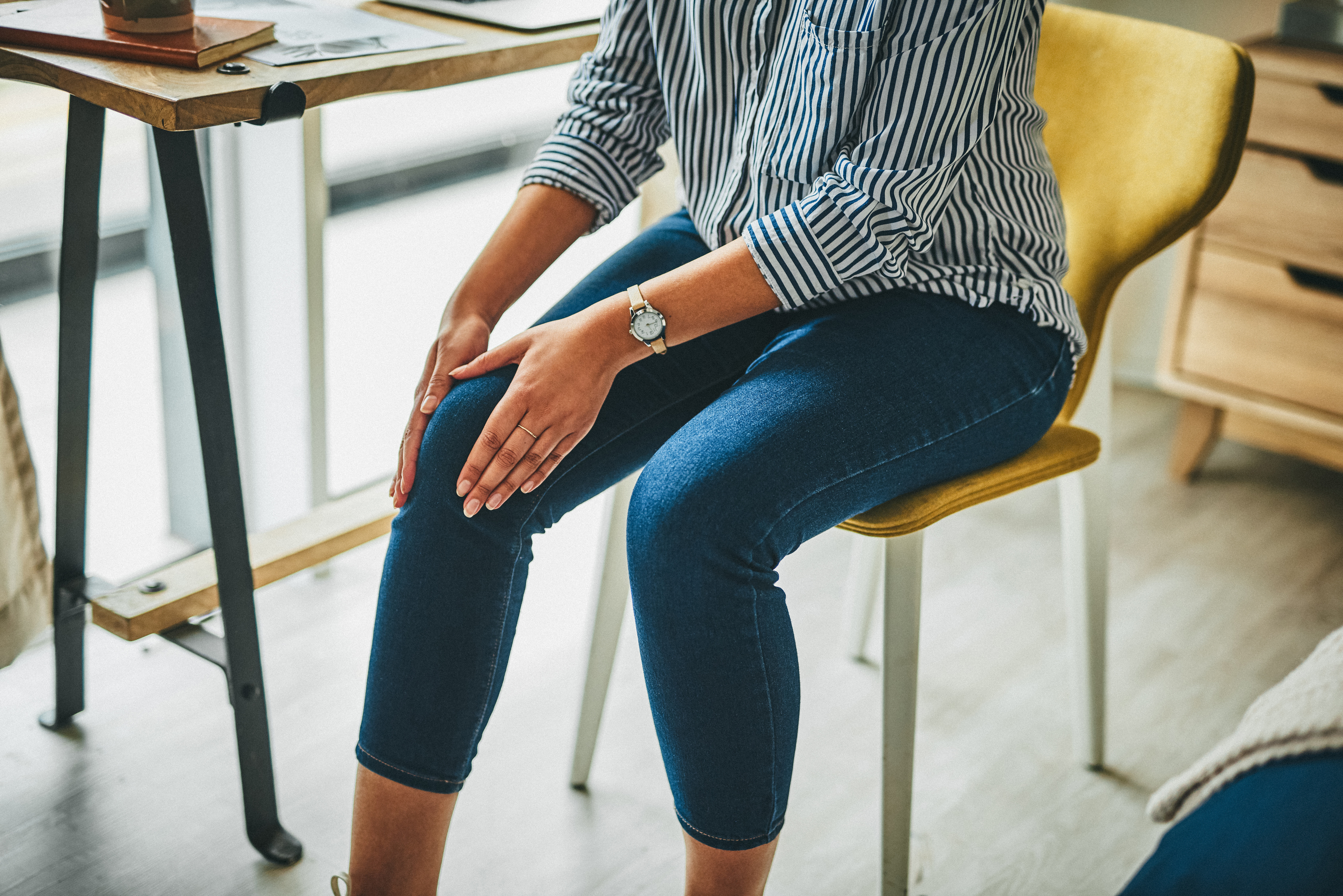 A person in a striped shirt and jeans sits on a chair indoors with their hands resting on their knees