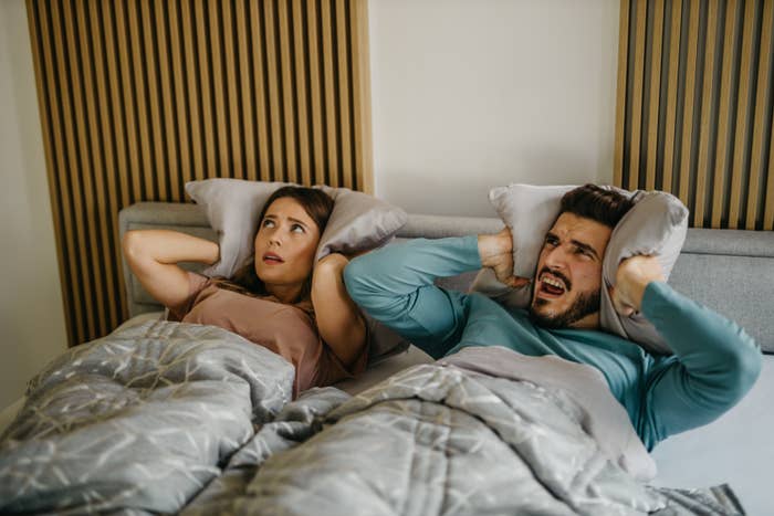 A man and woman lie in bed, both holding pillows over their ears and looking distressed as if disturbed by a loud noise