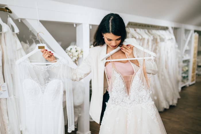 A woman is choosing between two wedding dresses, looking at one in each hand, while standing in a bridal shop