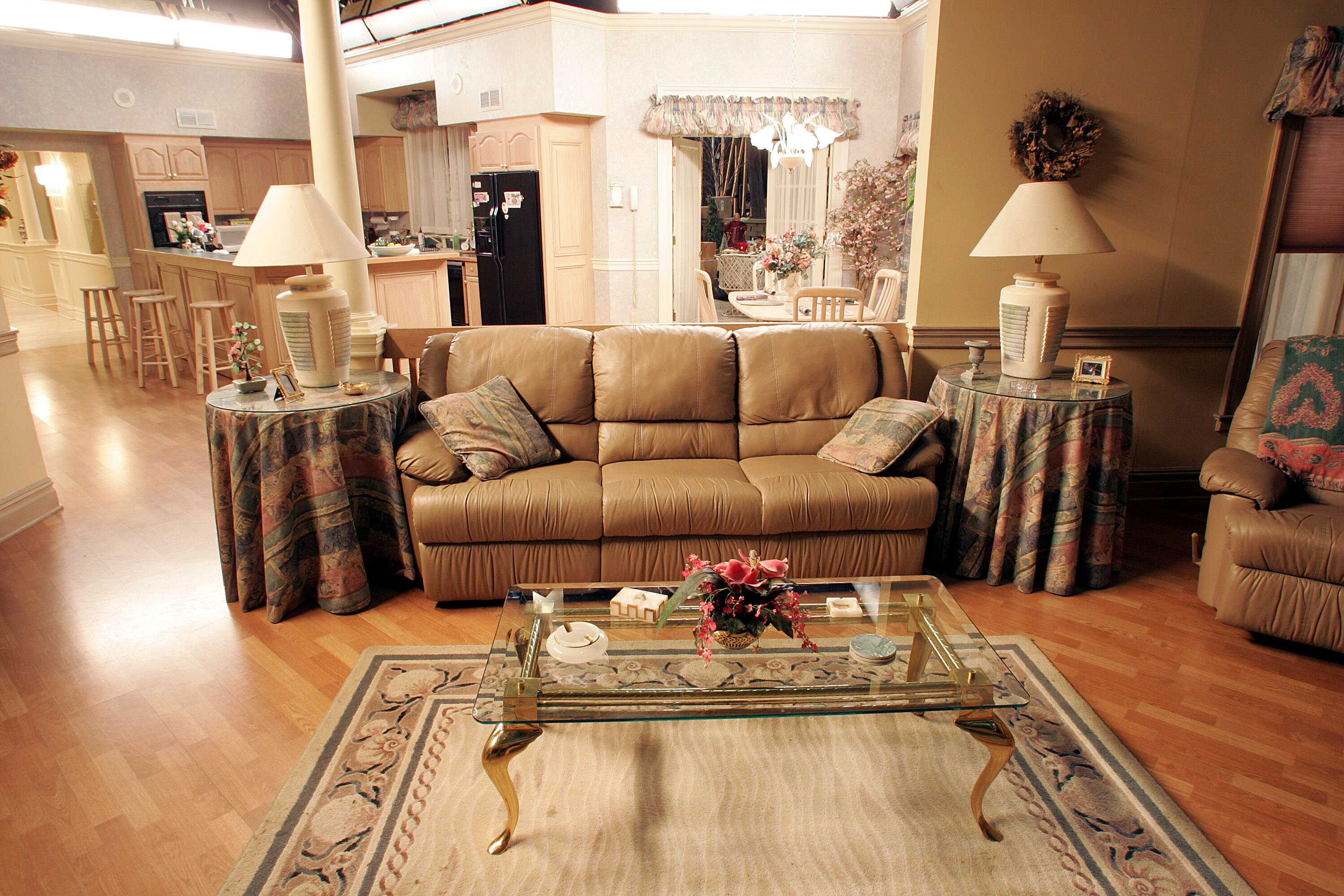 A cozy living room with a tan leather couch, glass coffee table, and matching side tables with lamps. Beyond, there is a view into the kitchen and dining area