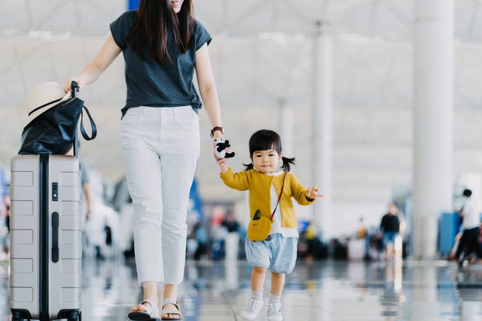 A woman holds the hand of a young girl, who is walking and holding a stuffed toy. They are in an airport, with the woman carrying a suitcase