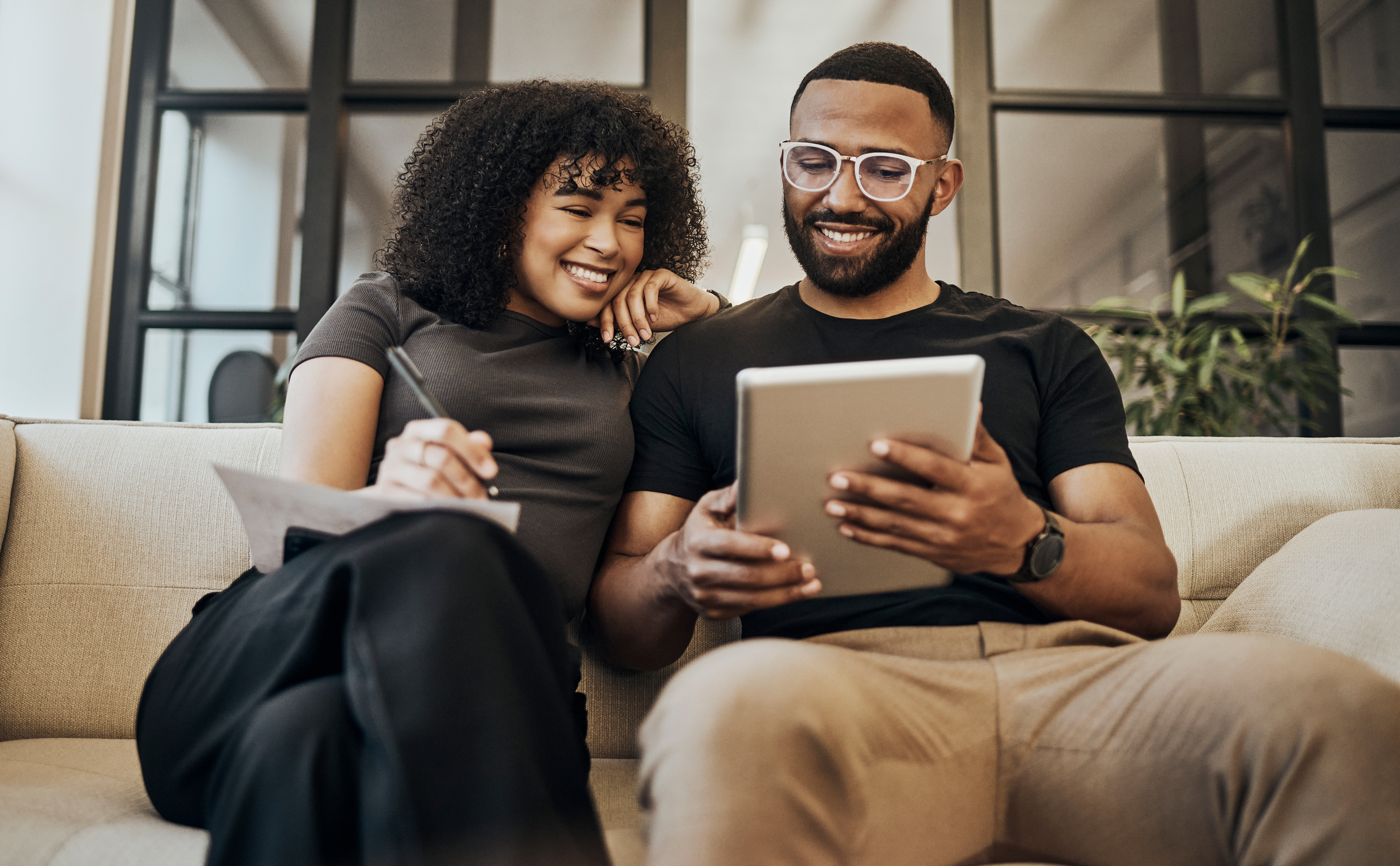A man and woman sit on a couch smiling and looking at a tablet. The woman holds a notebook and pen while the man wears glasses