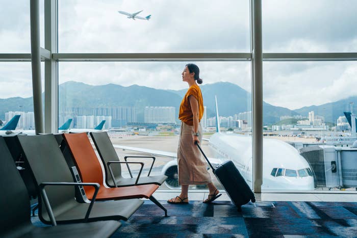 A woman walks through an airport terminal pulling a suitcase, with planes and mountains visible through large windows in the background