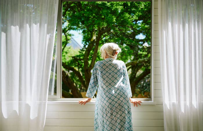 Elderly woman with silver hair and patterned robe stands at a window, looking out at a tree