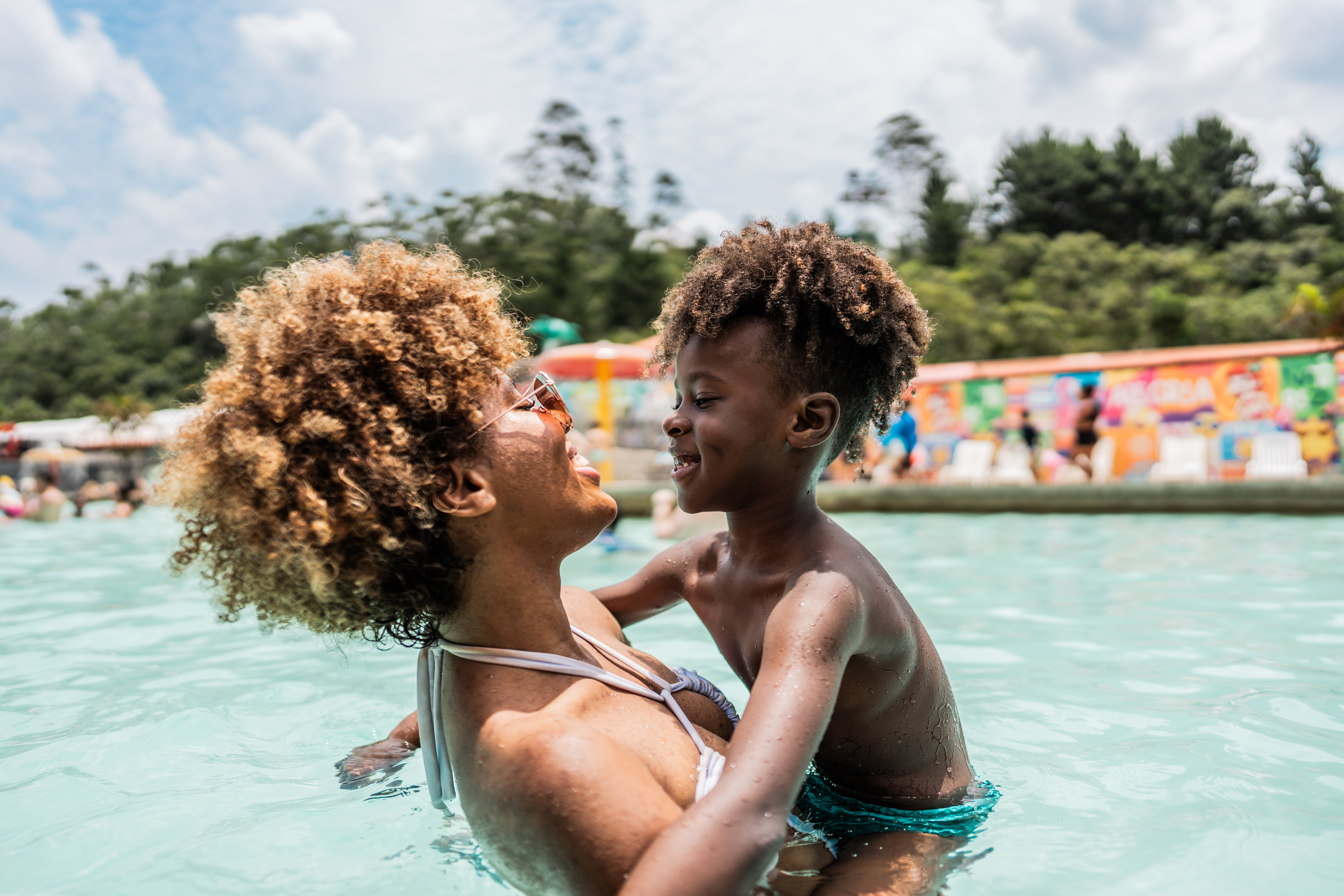 A parent and child are playing together in a swimming pool, smiling and enjoying their time under a cloudy sky