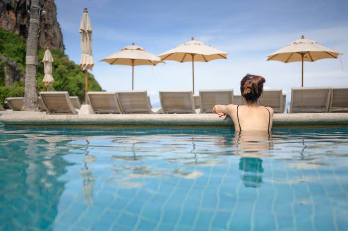 A person with their back facing the camera is relaxing in a hotel pool, looking at empty sun loungers and umbrellas on a sunny day