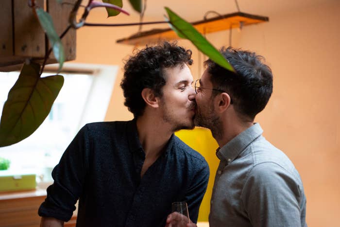 A couple, one with curly hair and the other with a beard and glasses, share a kiss in a cozy indoor setting. One person holds a drink