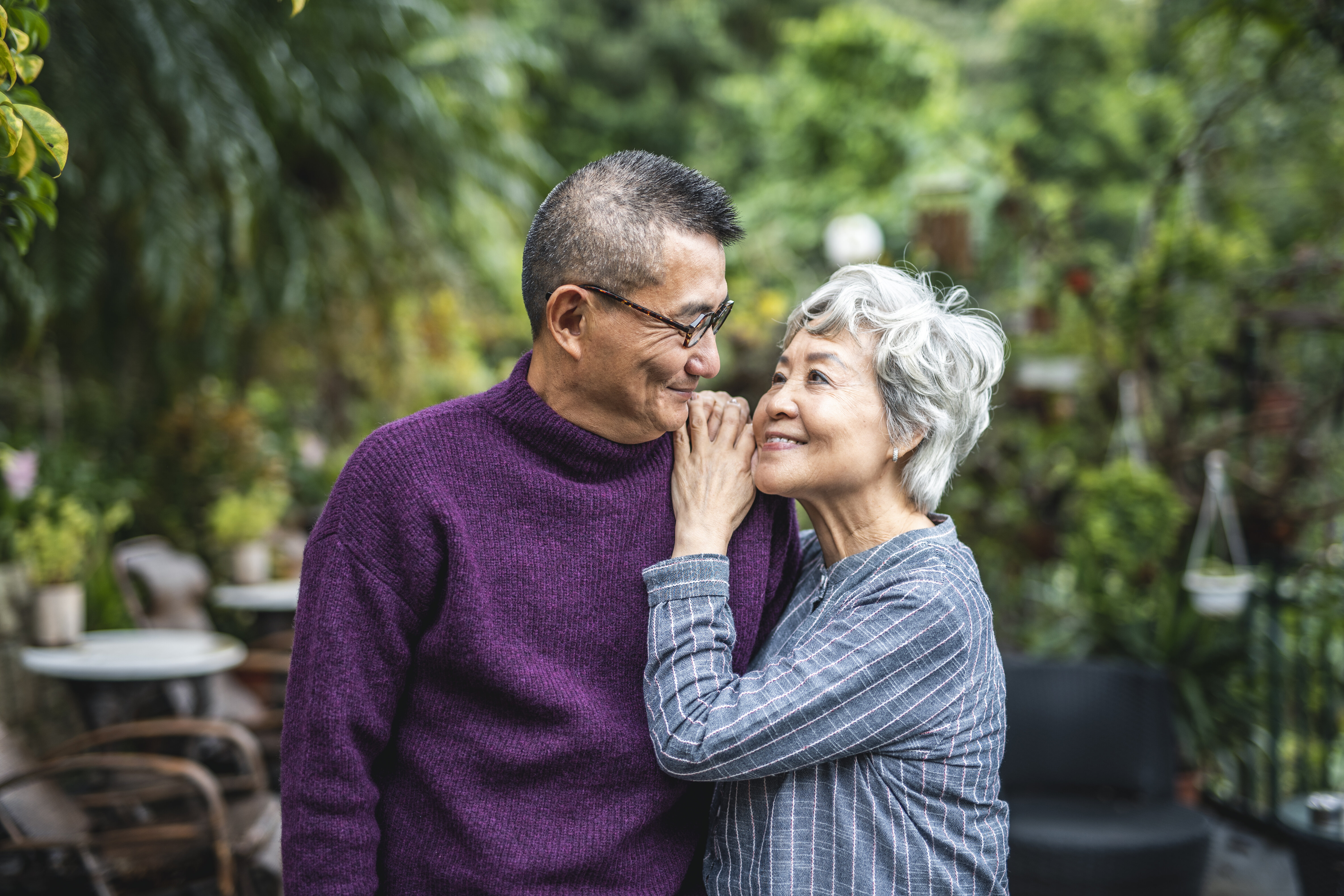 An elderly couple stands close in a garden, smiling affectionately at each other. The man wears a sweater, and the woman touches his shoulder gently