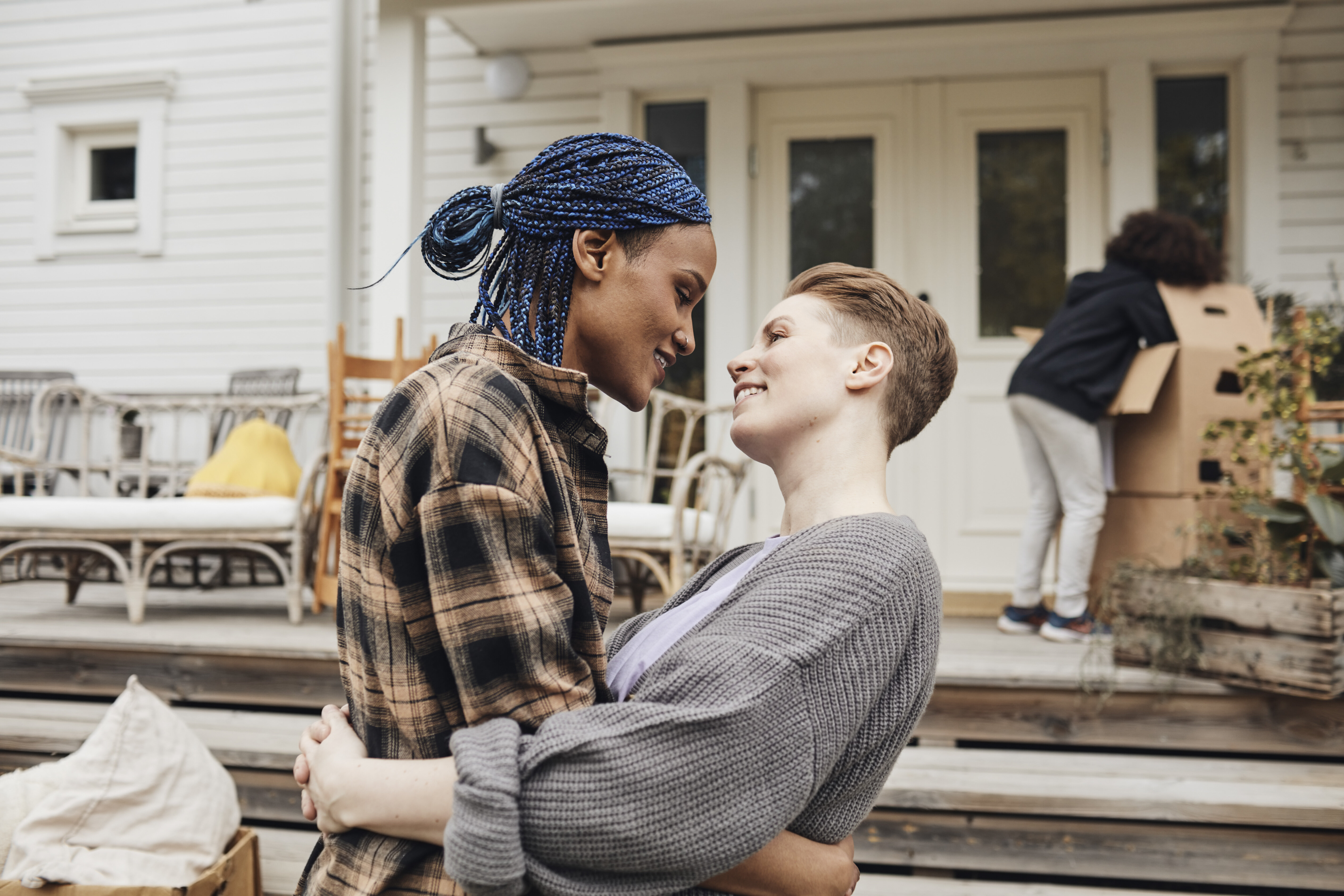 Intimate moment as two people embrace and smile at each other on a porch, moving boxes being unpacked in the background