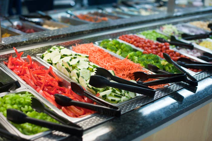 A salad bar with trays of various vegetables including bell peppers, cucumbers, shredded carrots, broccoli, cherry tomatoes, and celery