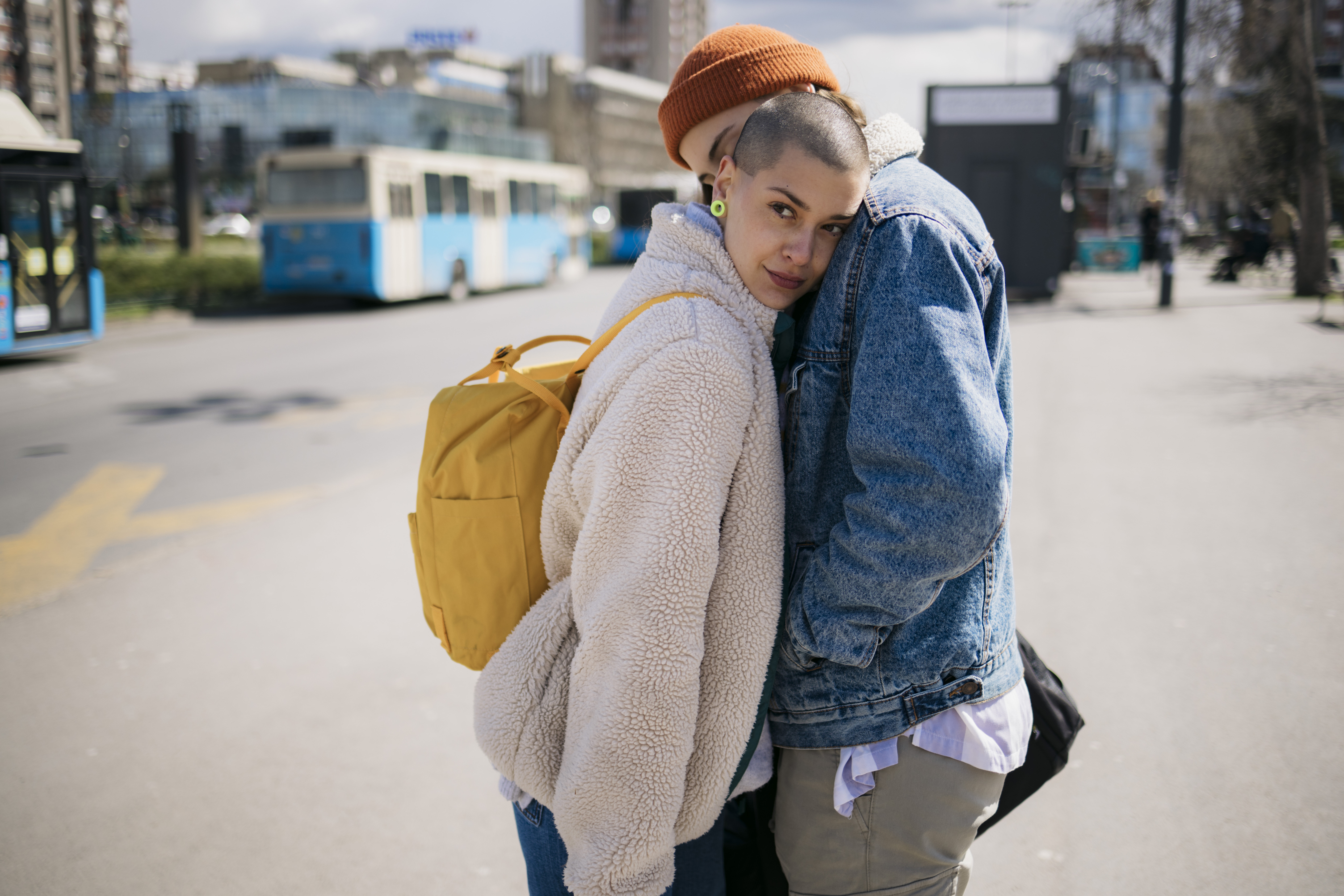 A person with a shaved head leans affectionately against another person in a denim jacket on a city street. Both have casual outfits; one has a yellow backpack
