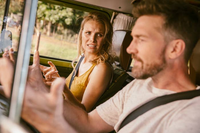 A woman and man sit inside a car, both wearing casual clothes. The woman looks upset and gestures towards the man, who is speaking and gesturing with his hands