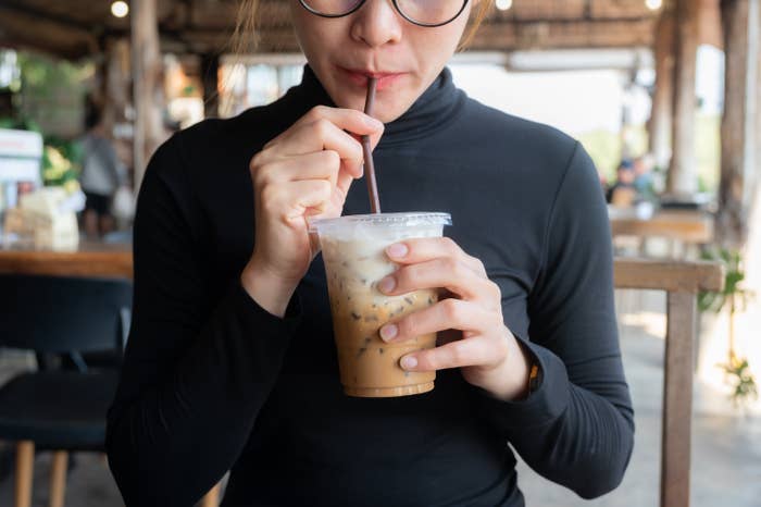A person wearing glasses and a turtleneck drinks iced coffee through a straw at a cafe