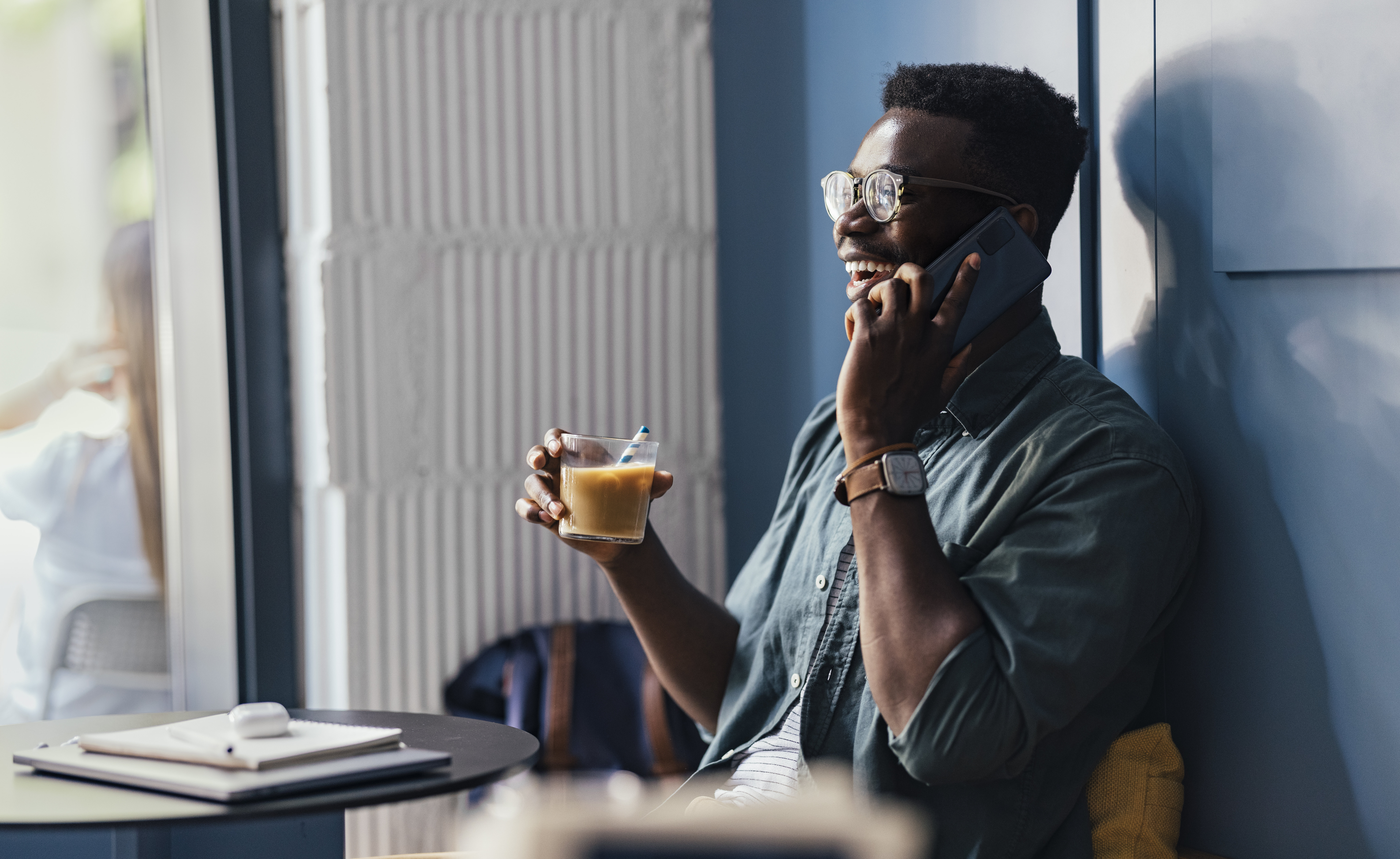 A person is laughing while talking on the phone and holding a drink in a cafe, with a notebook and pen on the table