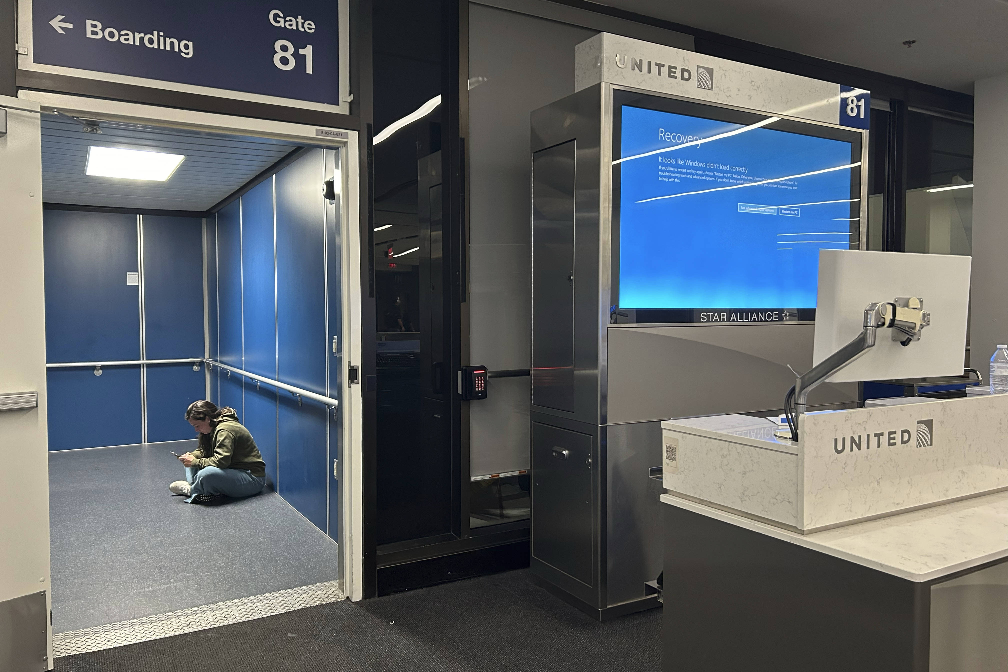 A person sits on the floor by the boarding gate 81 of United Airlines. A boarding sign and an empty customer service desk are visible
