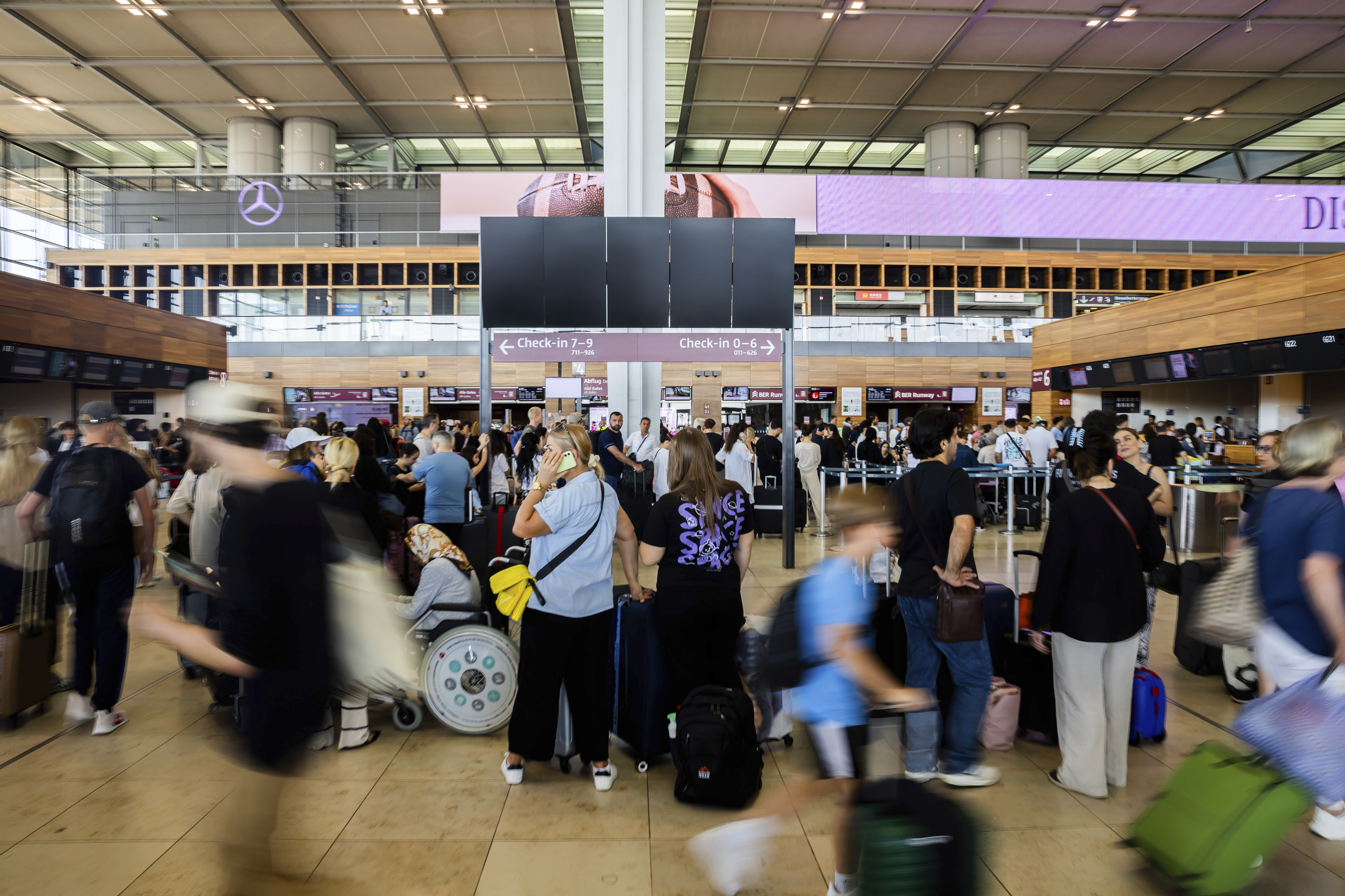 Busy airport terminal with numerous travelers moving through check-in areas. The background shows various check-in counters and digital screens displaying flight information