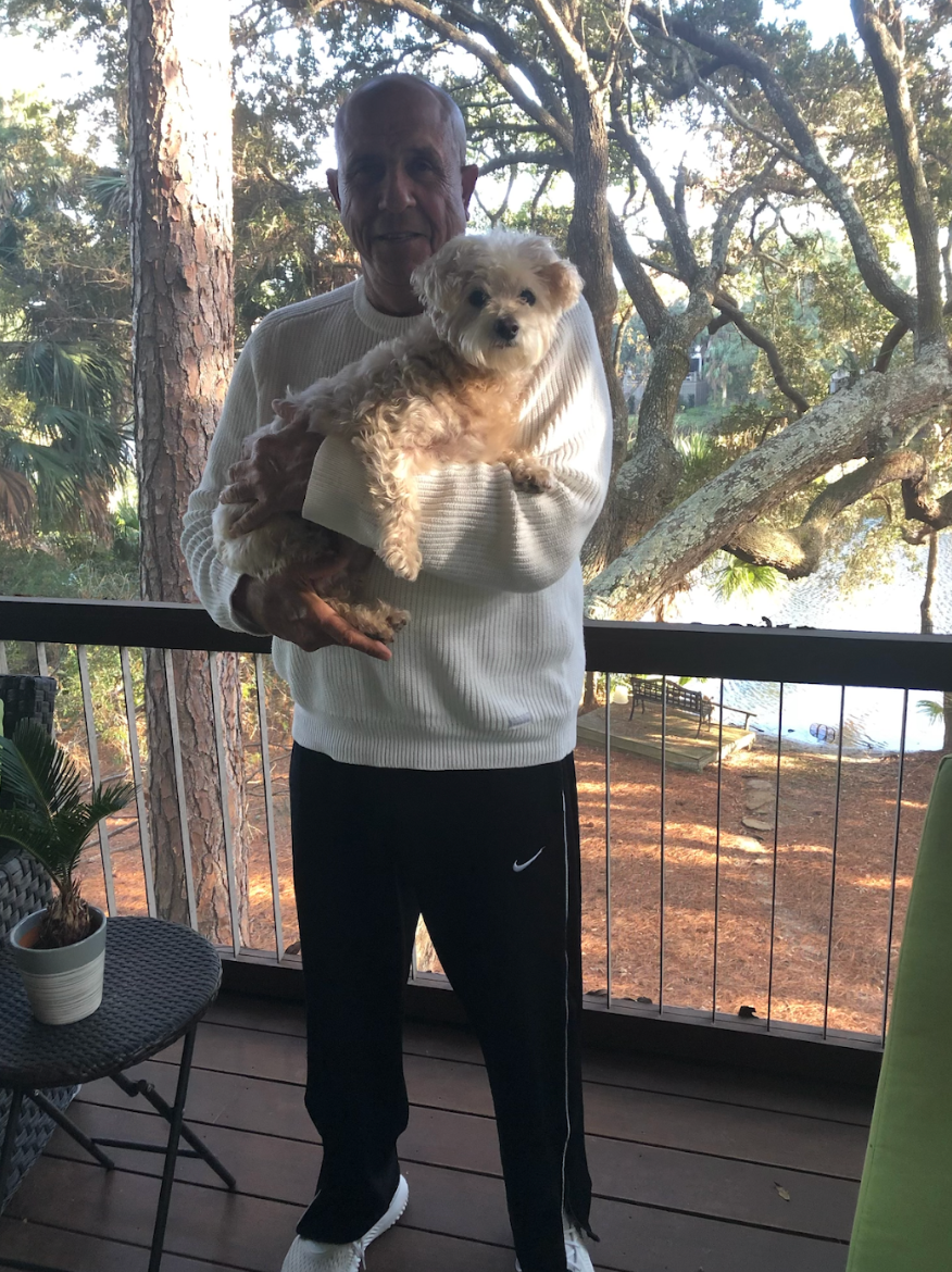 A person holding a fluffy dog on a deck with trees in the background. The person is wearing a sweater and pants