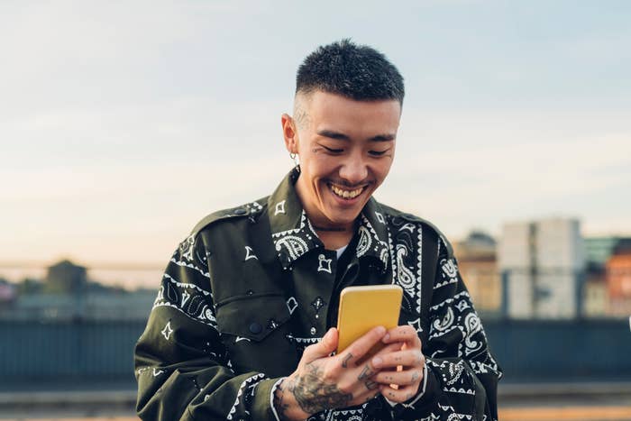 A smiling person with tattoos on their hands is looking at a yellow phone. They are wearing a bandana-patterned jacket. There are buildings blurred in the background