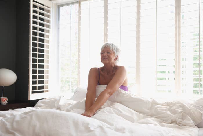 A woman with short gray hair stretches contentedly while sitting on a bed, bathed in natural light from the large windows behind her