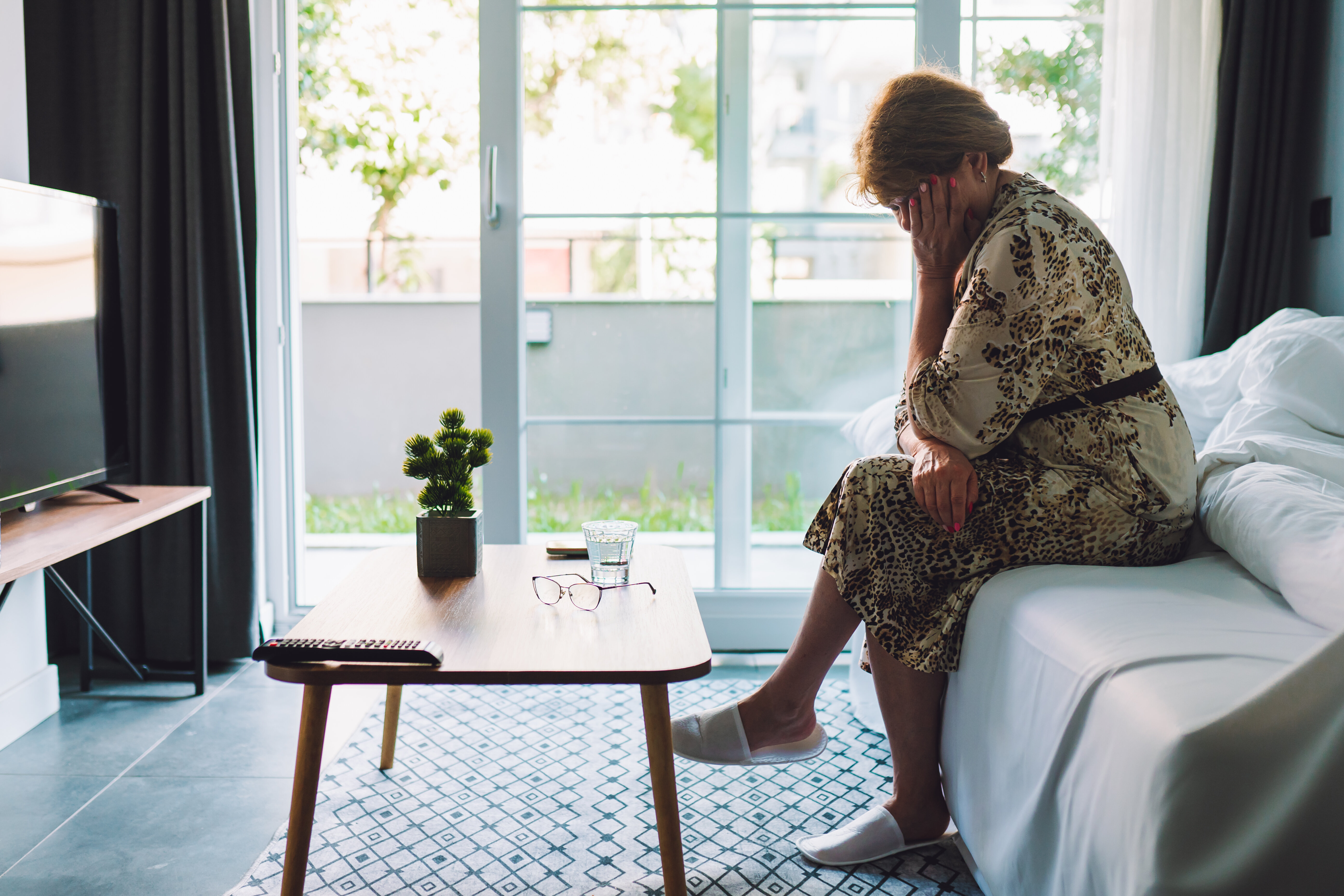 A person sits on a bed in a modern living room, head in hand, appearing distressed. Nearby are a table, a remote, a plant, a glass of water, and eyeglasses