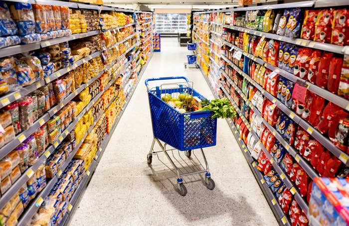 A grocery store aisle lined with various packaged food products and a shopping cart filled with fresh vegetables and fruits in the center