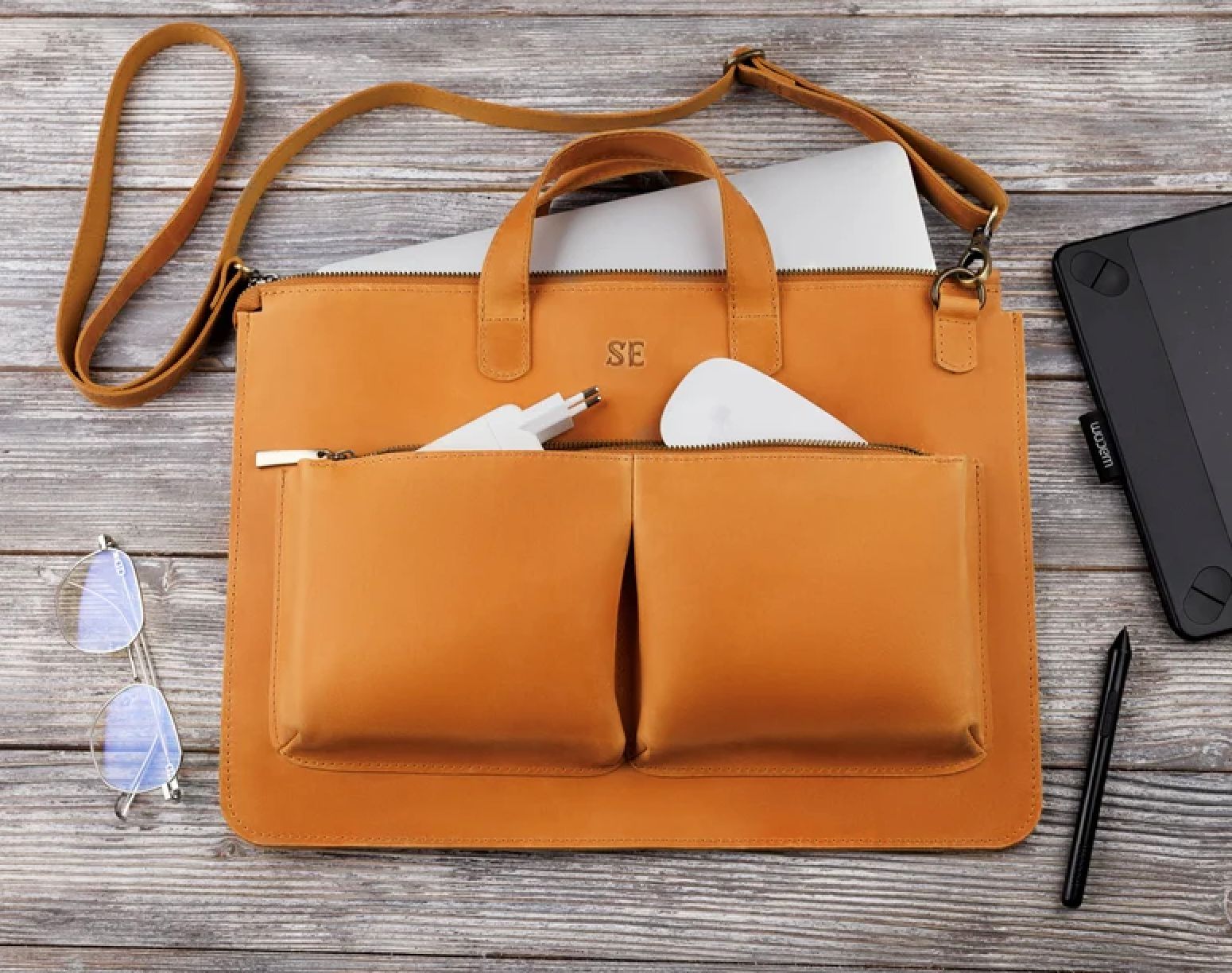 A leather laptop bag with two front pockets, containing a charger and a mouse. Accessories include glasses, a pen, a tablet, and a folded laptop inside the bag