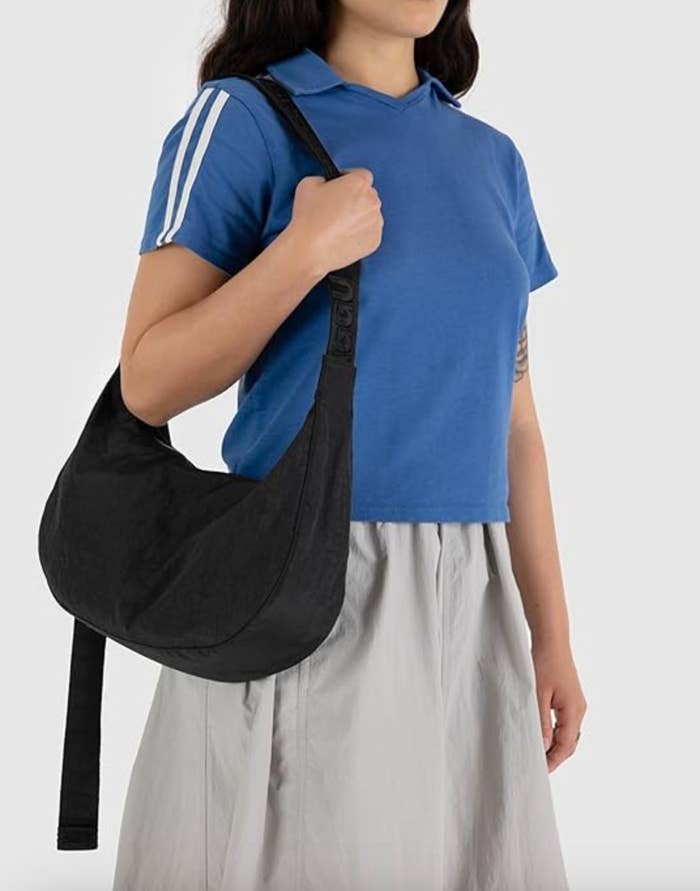 A person wearing a blue polo shirt and a gray skirt holds a black shoulder bag over their left shoulder