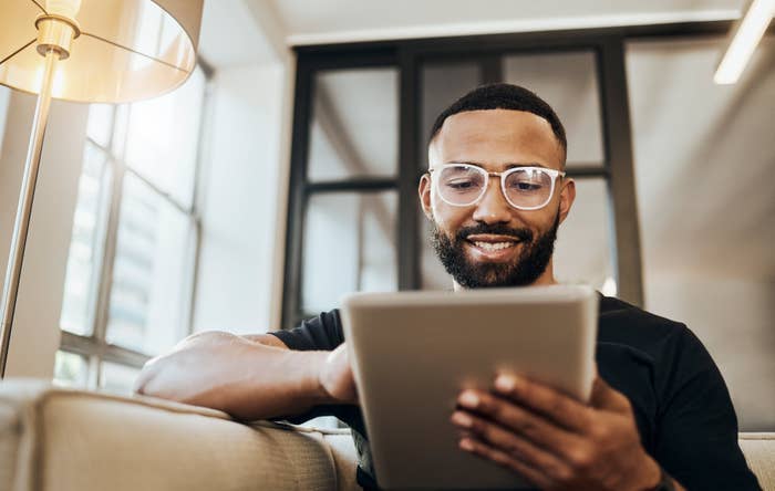 A man with glasses and a beard is sitting on a beige couch in a modern living room, smiling while using a tablet. He is near a lamp and large windows