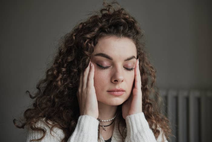 A woman with curly hair wearing a sweater and a pearl necklace has her eyes closed and holds her temples with both hands