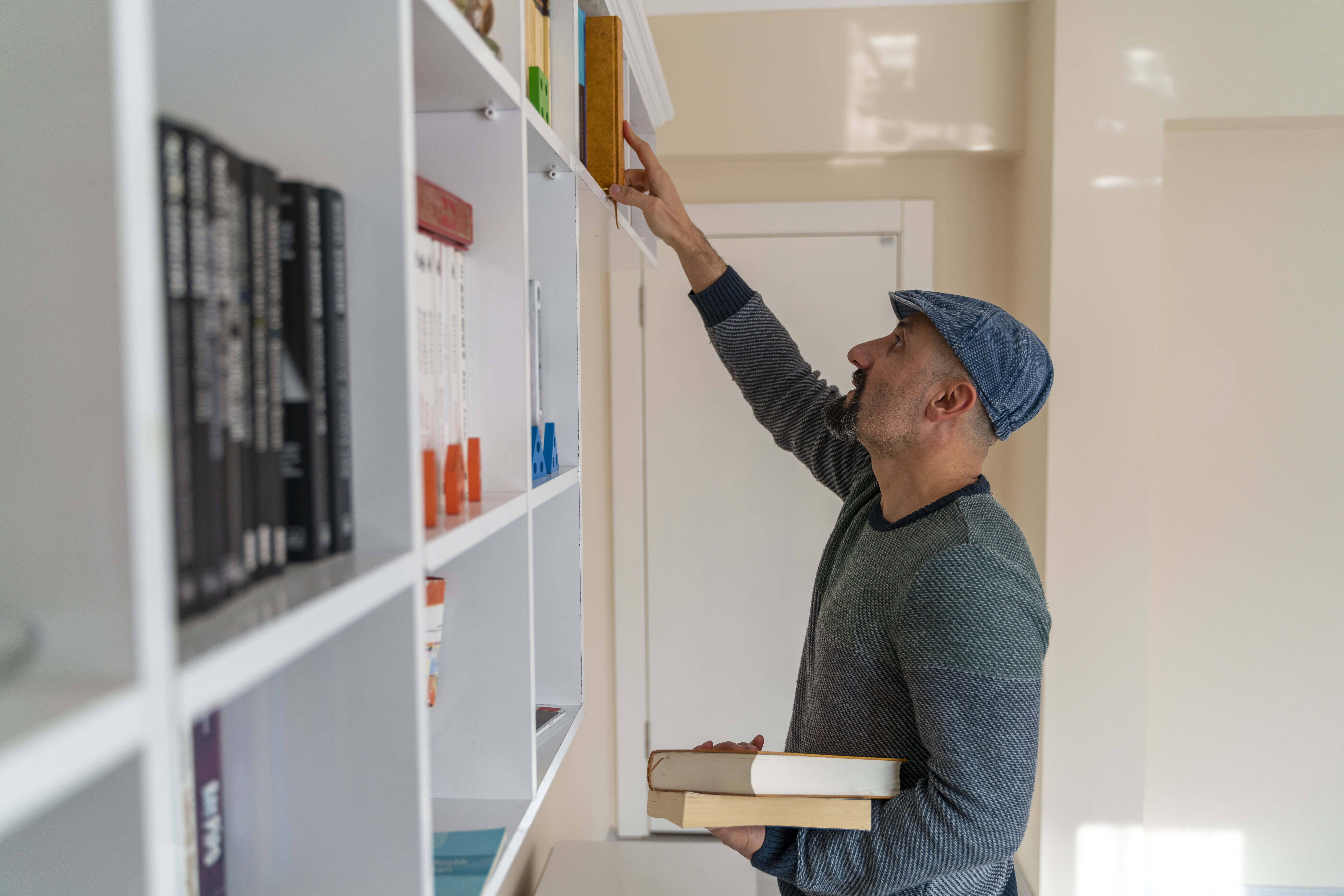 A man in a cap and sweater reaches for a book on a high shelf while holding two other books in his left hand. Shelves are filled with various books