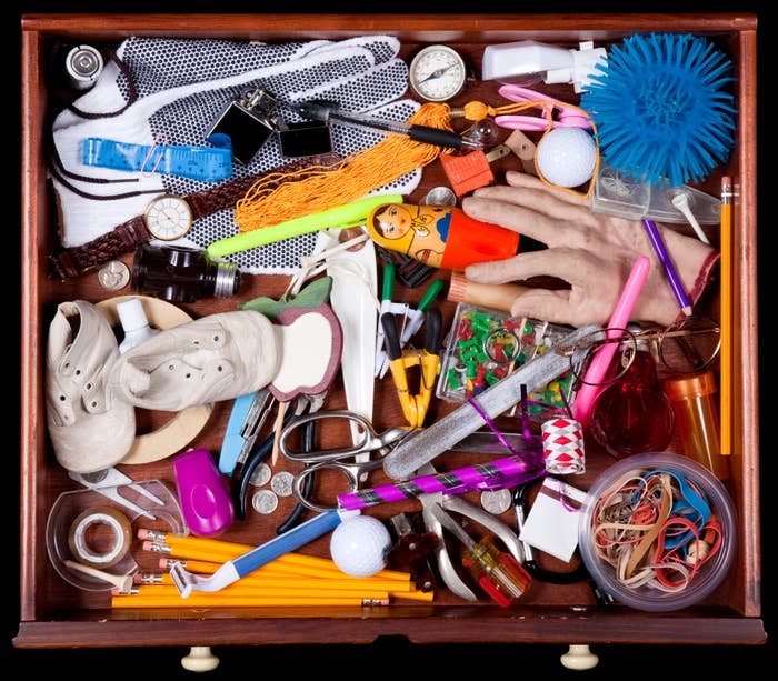 A cluttered drawer filled with various household items including a toy hand, golf balls, playing cards, rubber bands, coins, sunglasses, pens, and small tools