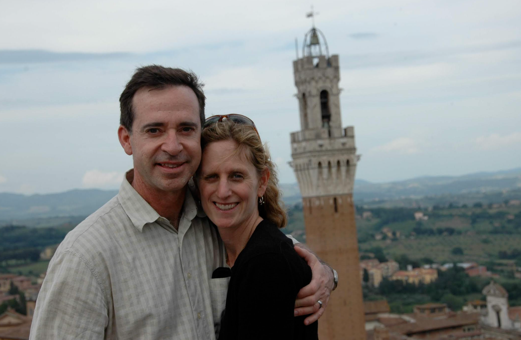 A couple smiles in front of a tall tower on a scenic hillside, enjoying the view
