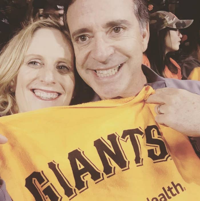 Two people smiling, holding up a &quot;Giants&quot; towel at a sports event. Crowds and branded gear are visible in the background