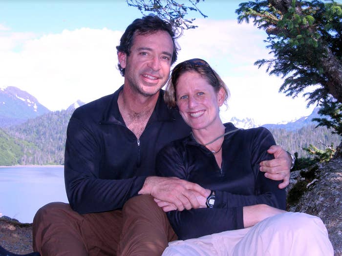A couple sits together, smiling, in an outdoor setting with mountains and water in the background