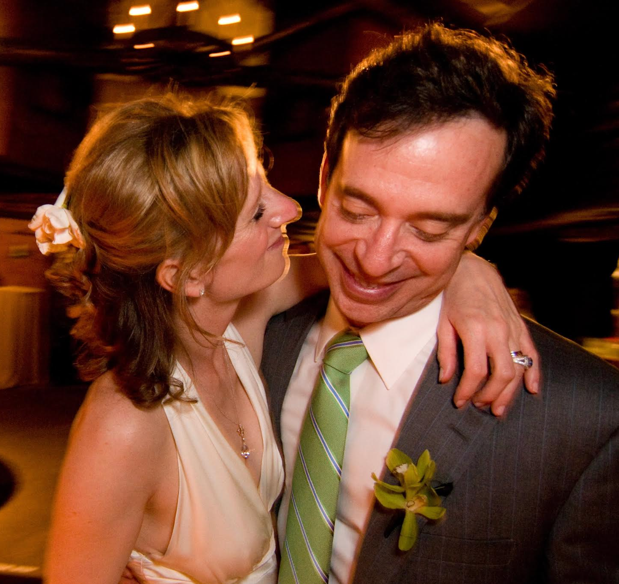 A woman in a sleeveless dress with a flower in her hair shares a joyful moment with a man in a suit and striped tie adorned with a green boutonniere