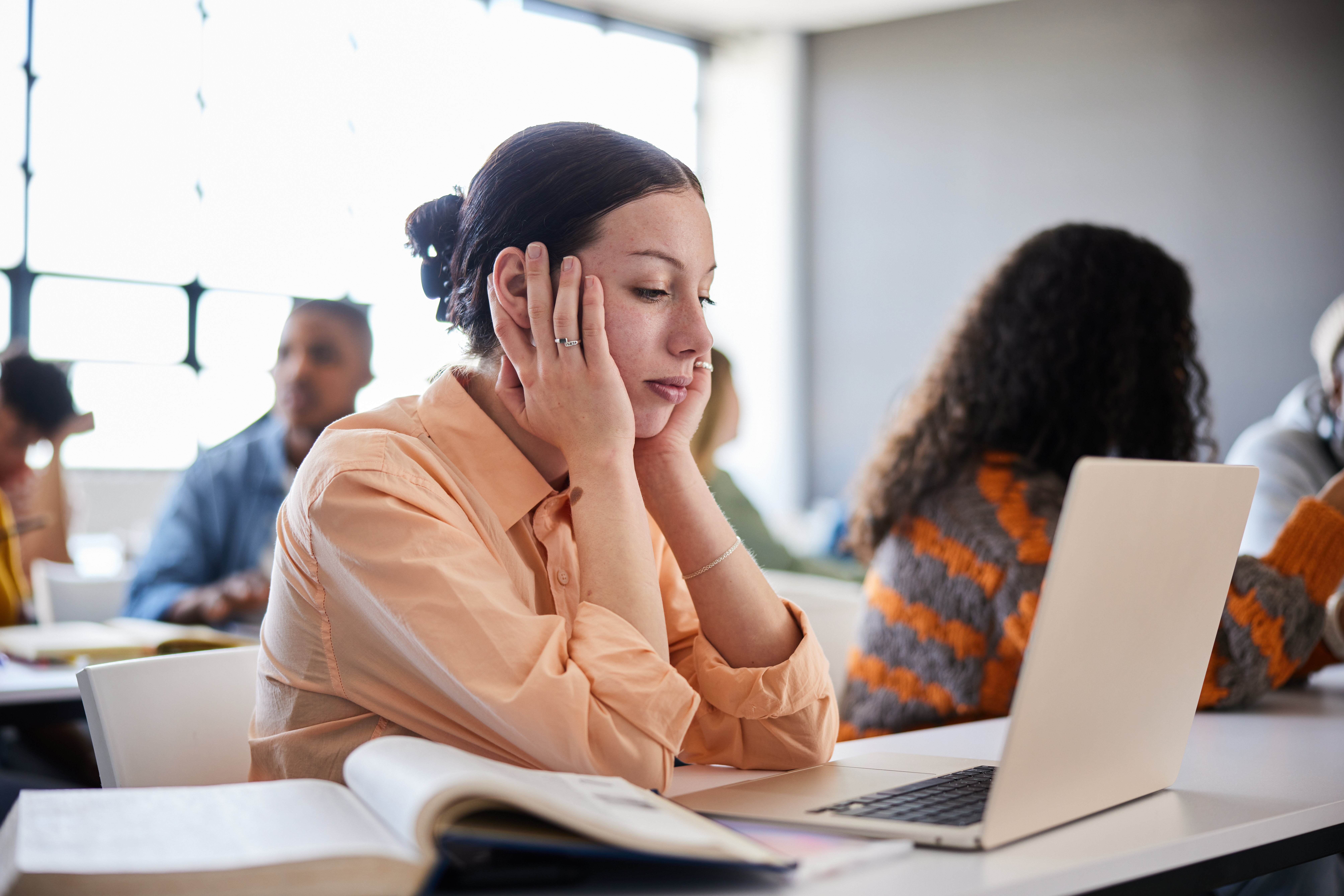 A woman sits at a desk, resting her face in her hands while looking at a laptop. An open book is in front of her. Other people are working in the background