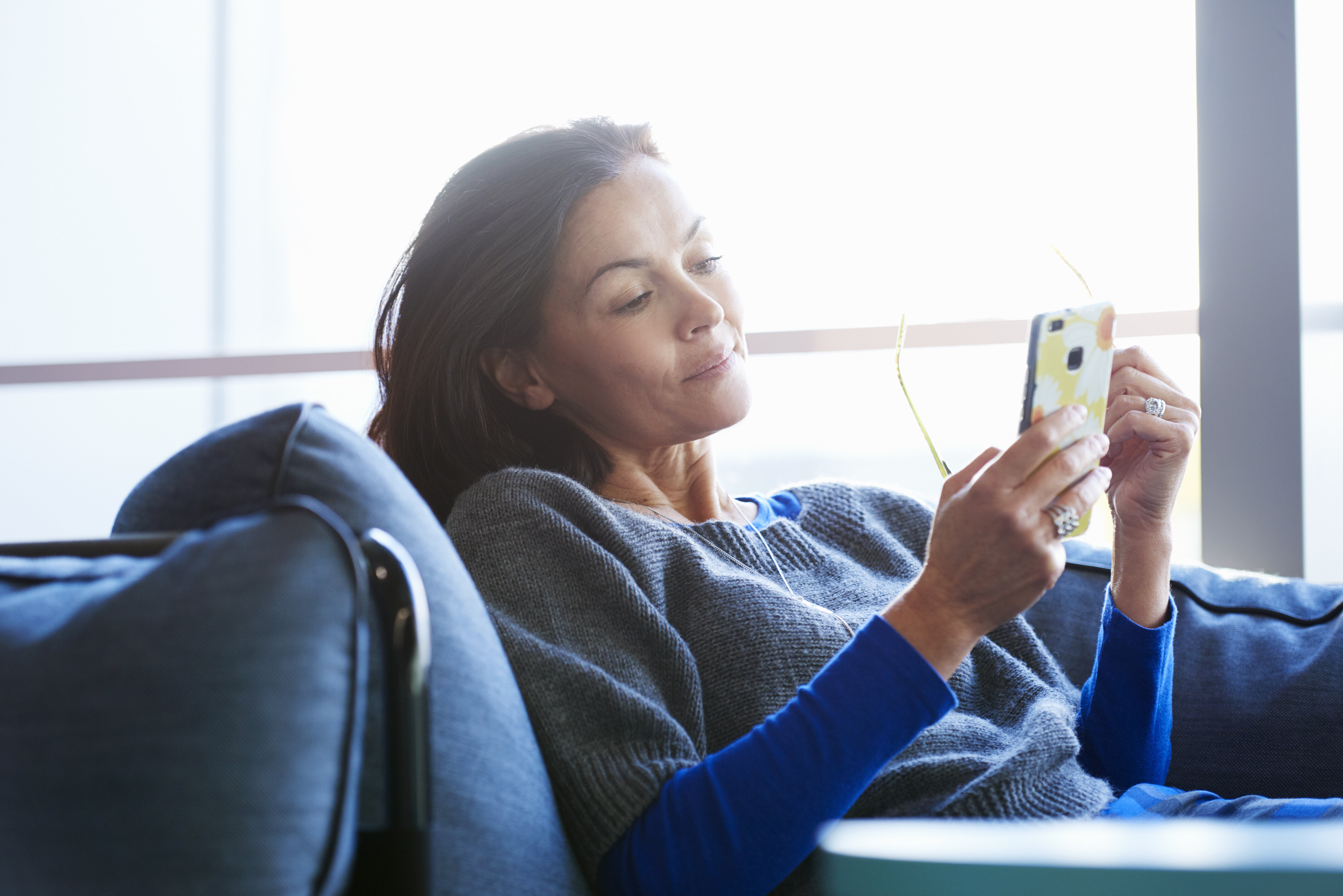 A woman is relaxing on a couch, looking at her phone, with reading glasses in her hand
