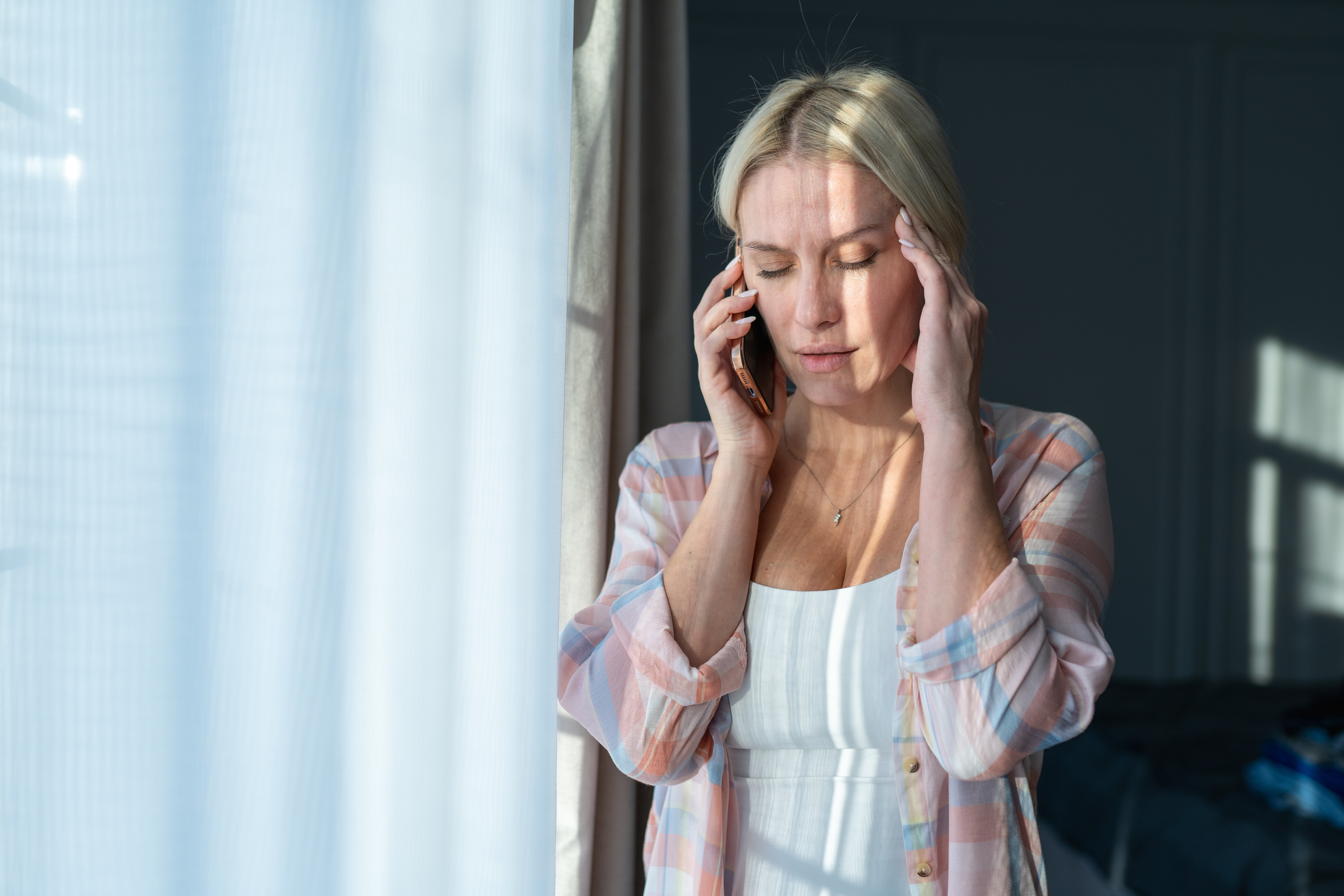 Woman in a room holding her head with one hand while talking on the phone. The sunlight streams in through the window blinds