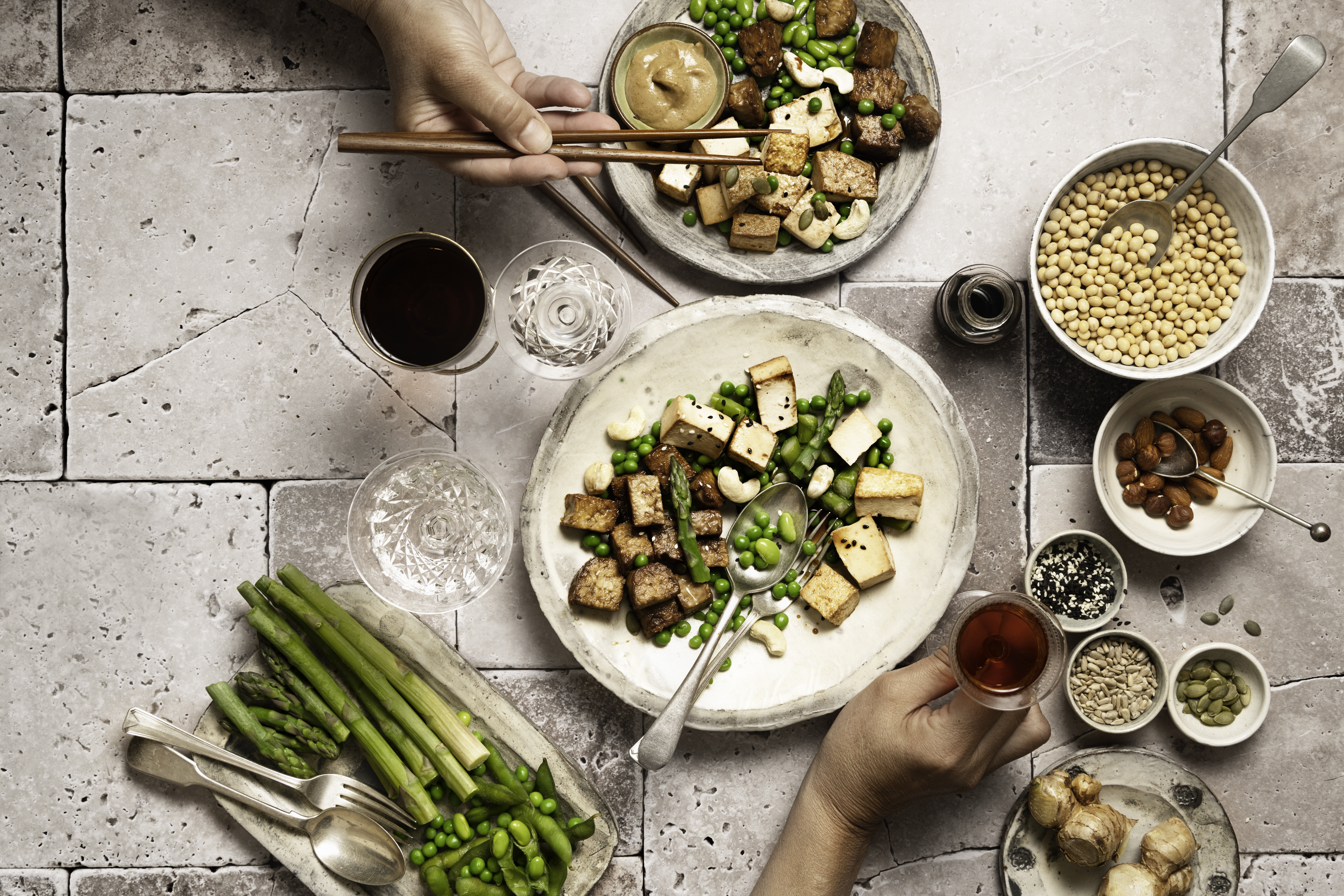 Hands arranging a healthy meal with tofu, vegetables, grains, and sauces on a rustic tiled surface. Bowls with green beans, chickpeas, almonds, and seeds are visible