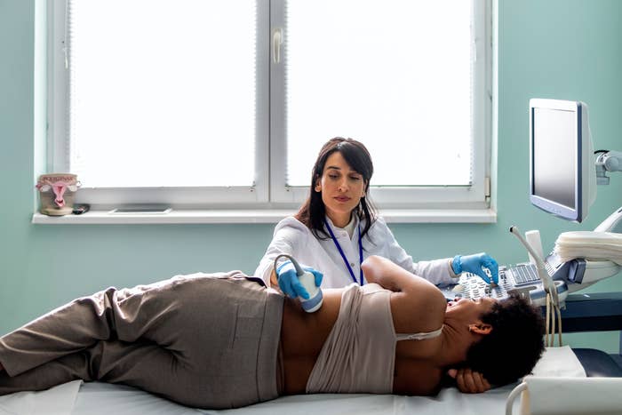 A medical professional performs an ultrasound on a patient lying on their side in a clinical setting. The patient is wearing a hospital gown