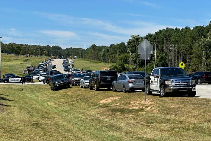 Numerous police vehicles line a rural road, with a backdrop of trees and grassy areas. This is a scene of a significant police response