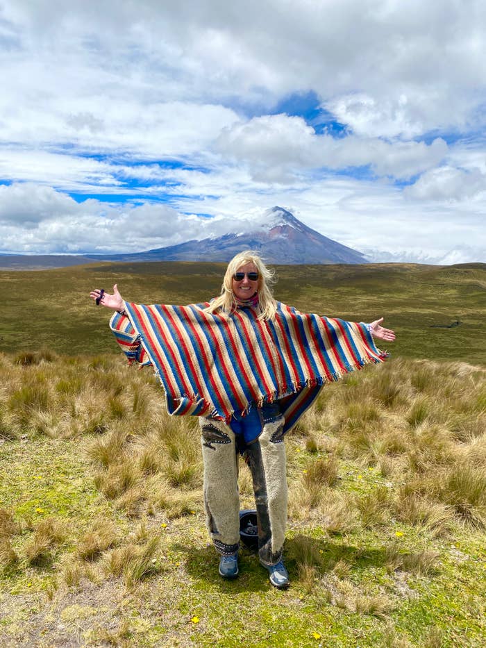 Person standing outdoors with open arms, wearing sunglasses, and a poncho with multicolored stripes. A mountain is visible in the background