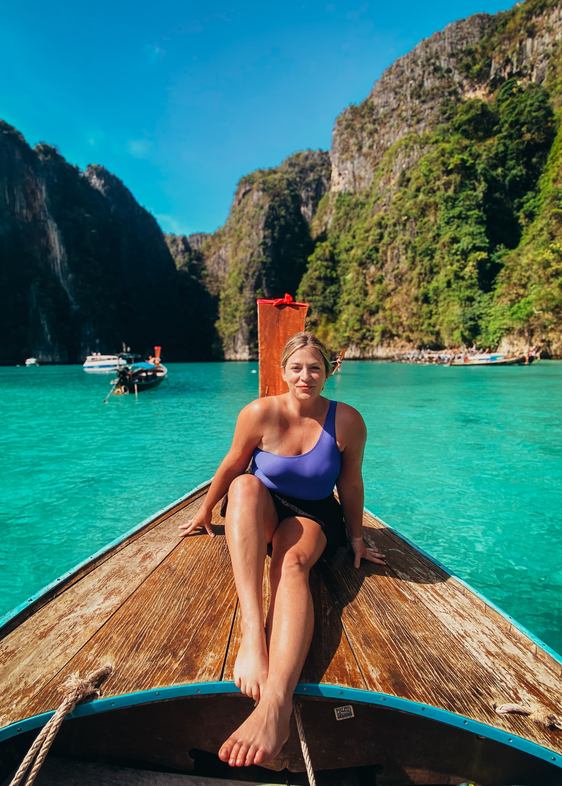 Woman in a swimsuit sits on the bow of a boat in a picturesque tropical lagoon with clear water and lush green cliffs in the background