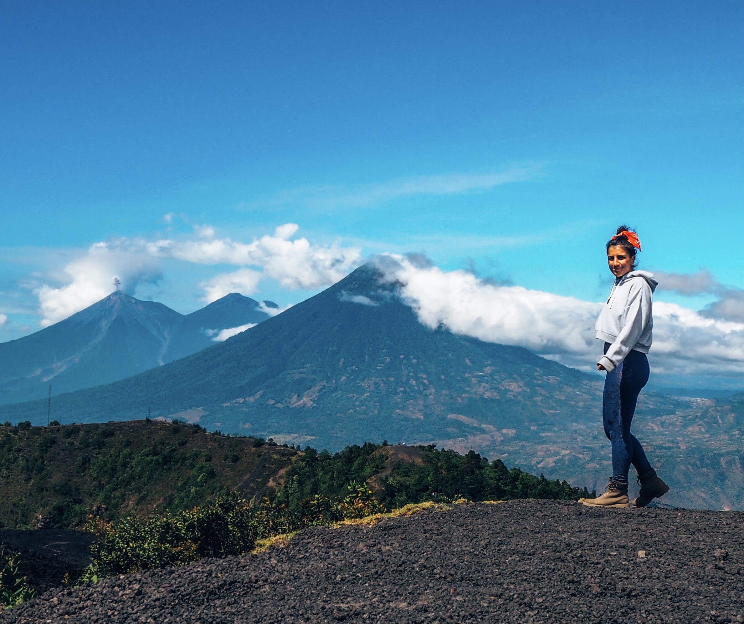 Woman in a hoodie and jeans standing on a mountain, with scenic view of cloud-covered volcanoes in the background