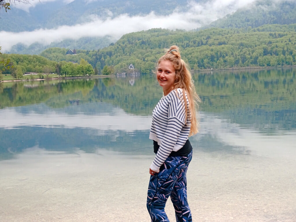 A woman with long hair in a topknot and wearing a striped sweater and patterned pants stands by a lake with mountains and fog in the background